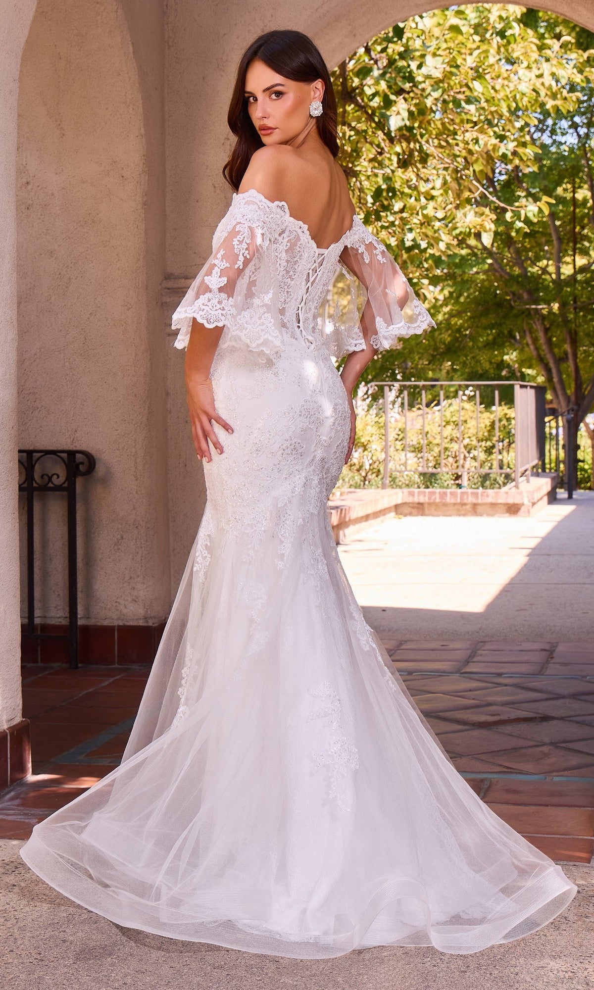 A woman wearing the Ladivine CD659W long wedding dress, featuring an off-shoulder lace design with a fitted bodice and flared mermaid skirt, stands outdoors near a stone archway and lush greenery.